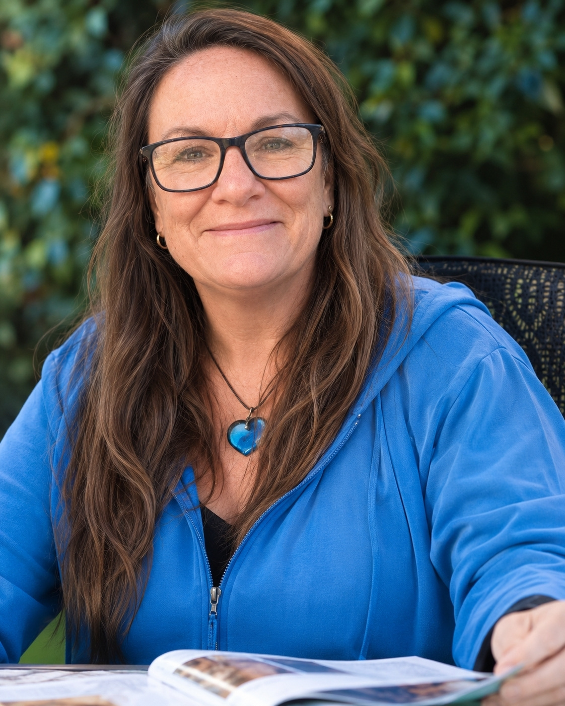 Friendly outdoor portrait of a woman with glasses and long brown hair, wearing a blue jacket and blue heart pendant while reviewing a magazine